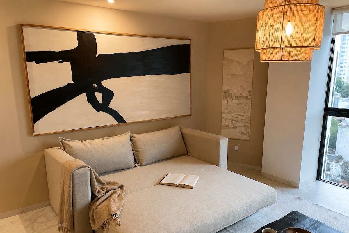Una sala de estar moderna con un cuadro abstracto grande, un sofá con un libro, una mesa de centro con un tazón y una taza, y una lámpara colgante.