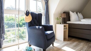 Une chambre avec une grande fenêtre, un fauteuil bleu foncé, une table de chevet avec une lampe et un lit avec des oreillers blancs.