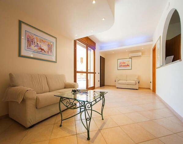 A spacious living room with a beige sofa, a glass coffee table, and two framed artworks on the walls.