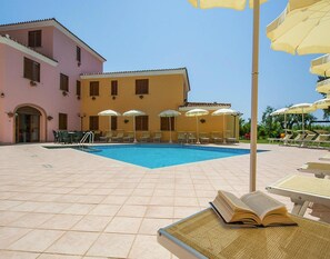 A pool area with lounge chairs and umbrellas, a building with a pink facade, and a clear blue sky. - Apartment in Sos Alinos Near Cala Liberotto (Orosei)