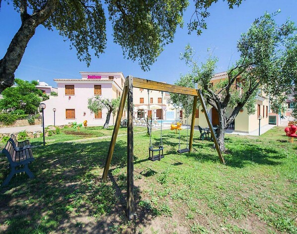 A playground with swings and a bench, surrounded by greenery and buildings in the background. - Apartment in Sos Alinos Near Cala Liberotto (Orosei)