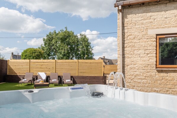 A hot tub with lounge chairs, a wooden fence, and a stone building.