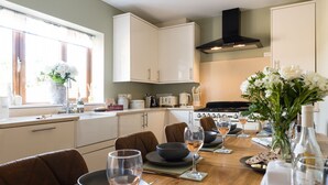 A modern kitchen with white cabinets, a black range hood, and a dining area with a wooden table set for four. - Incredible dog friendly holiday home in the Cotswolds with a hot tub - Puddleduck House (Bourton-on-the-Water)