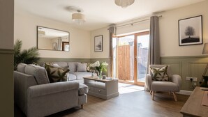 A living room with a grey sofa, a coffee table, and a chair. There is a wooden floor, a large window with curtains, and a chandelier. - Incredible dog friendly holiday home in the Cotswolds with a hot tub - Puddleduck House (Bourton-on-the-Water)