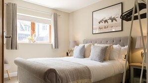 A bedroom with a large bed, a window with curtains, a bedside table with a lamp, and a framed picture on the wall. - Incredible dog friendly holiday home in the Cotswolds with a hot tub - Puddleduck House (Bourton-on-the-Water)