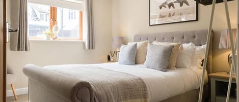 A bedroom with a large bed, a window with curtains, a bedside table with a lamp, and a framed picture on the wall.