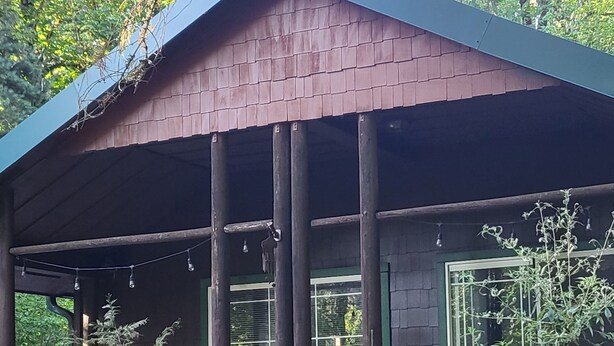 A rustic wooden cabin with a covered porch, a fenced outdoor area, and a view of the surrounding greenery. - Private enchanting cabin on the river  (McKenzie Bridge)