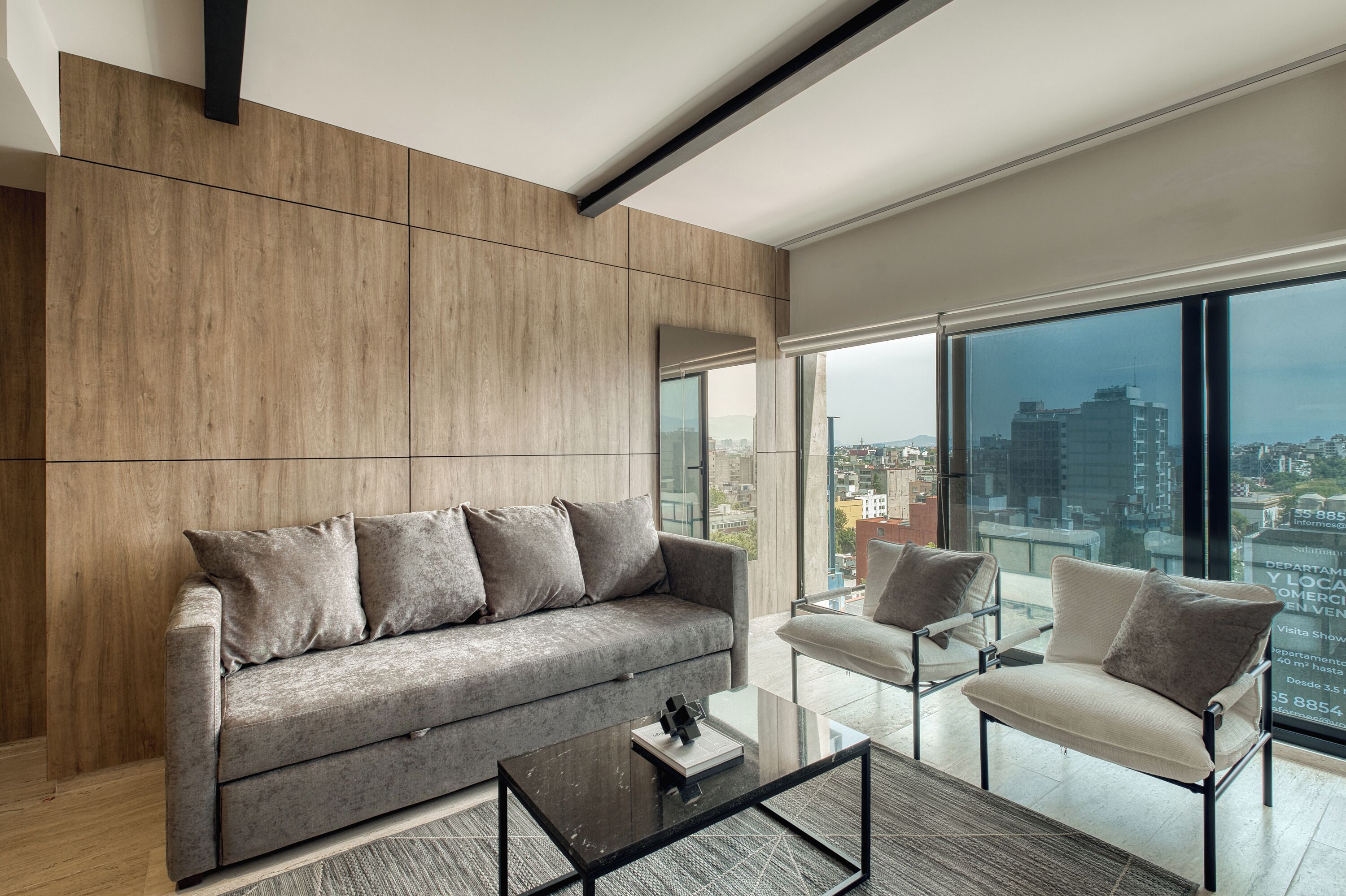 Ein modernes Wohnzimmer mit einem grauen Sofa, einem Glastisch und einem Sessel. Große Fenster bieten Blick auf die Stadt.