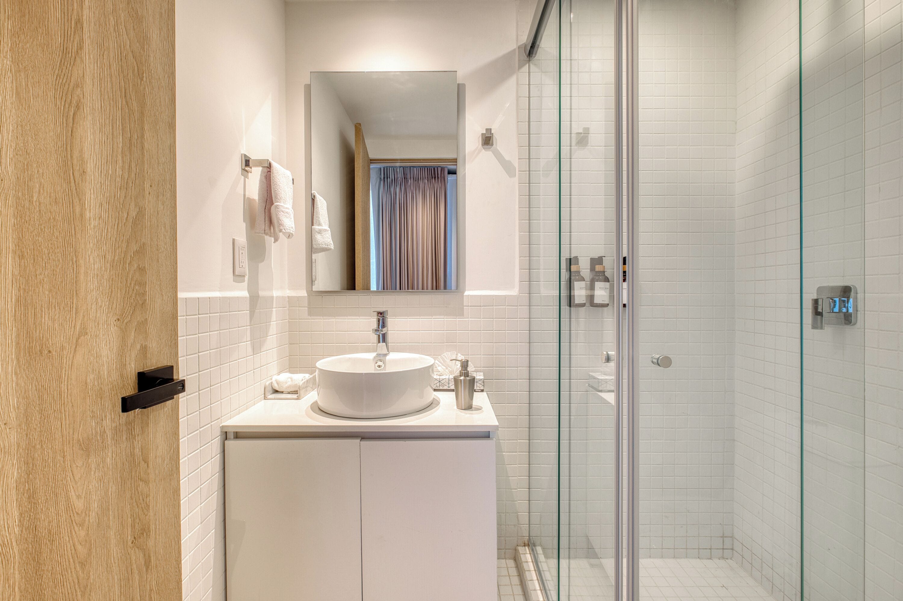 Een moderne badkamer met een douchecabine van glas, een wit wastafelmeubel met spiegel erboven en een houten deur.