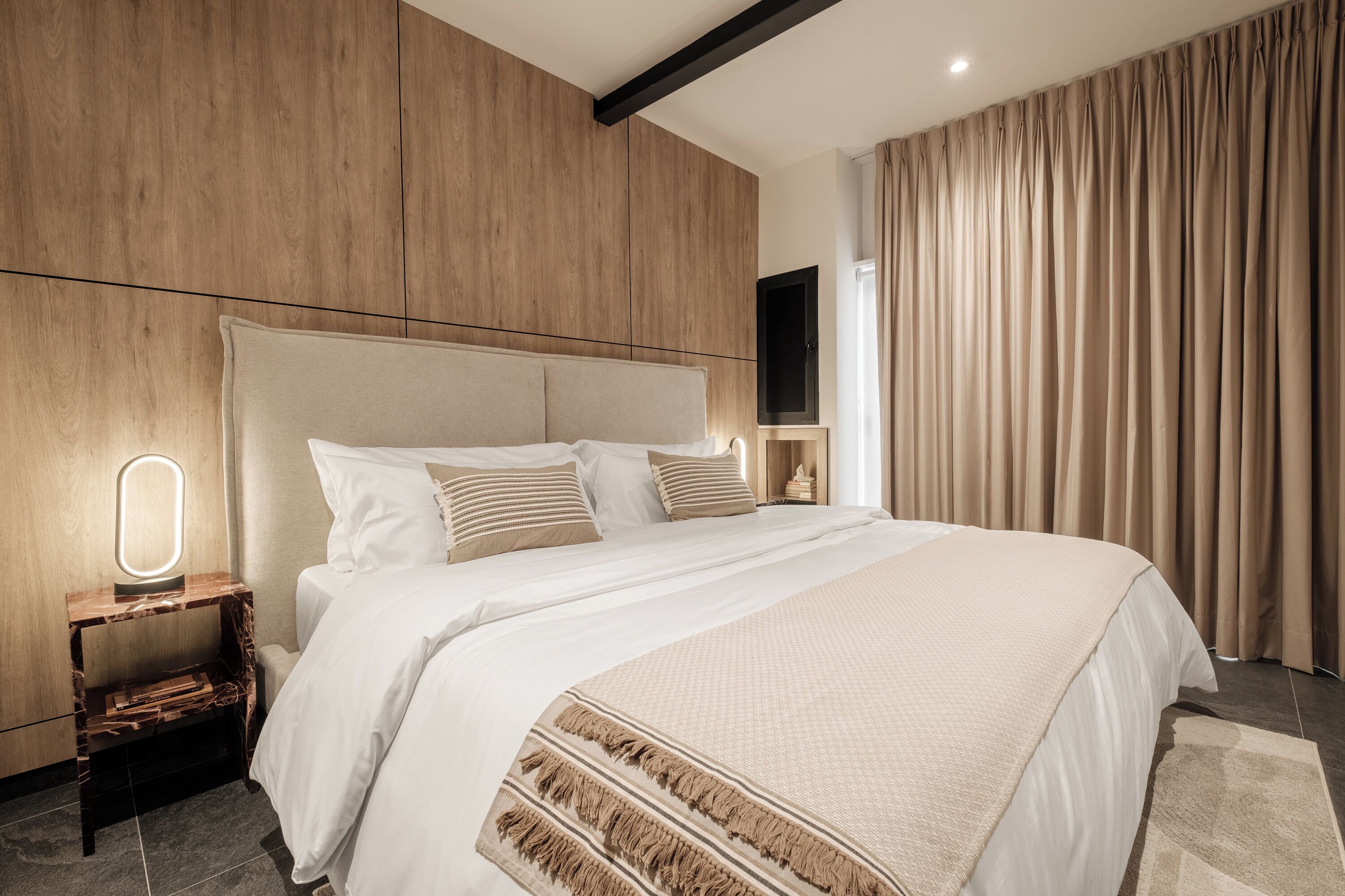 A modern hotel room with a large bed, a bedside table with a mirror, a flat-screen TV, and a large window with curtains.