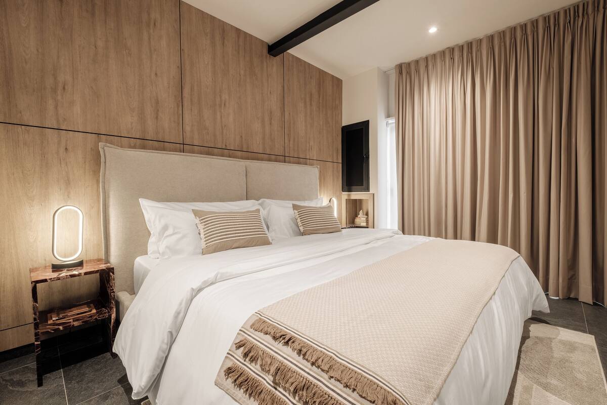 A modern hotel room with a large bed, a bedside table with a mirror, a flat-screen TV, and a large window with curtains.