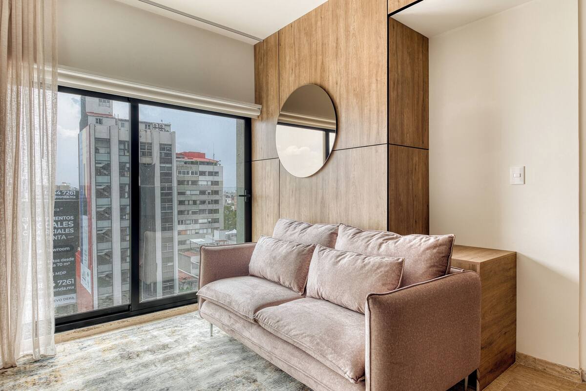 A modern living room with a large window, a round mirror, and a wooden accent wall.