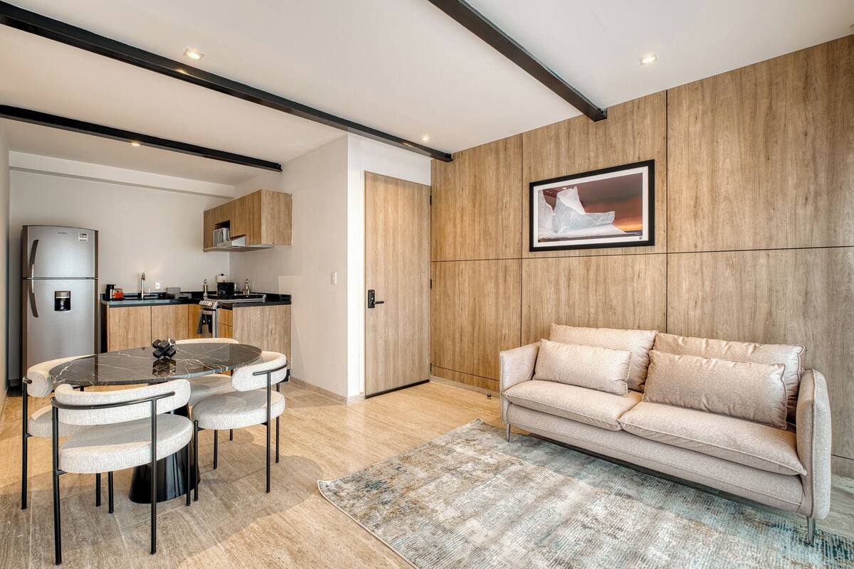 A modern kitchen with a built-in refrigerator, a dining area with a round table and chairs, and a beige sofa in a living area with a wooden accent wall and a framed picture.