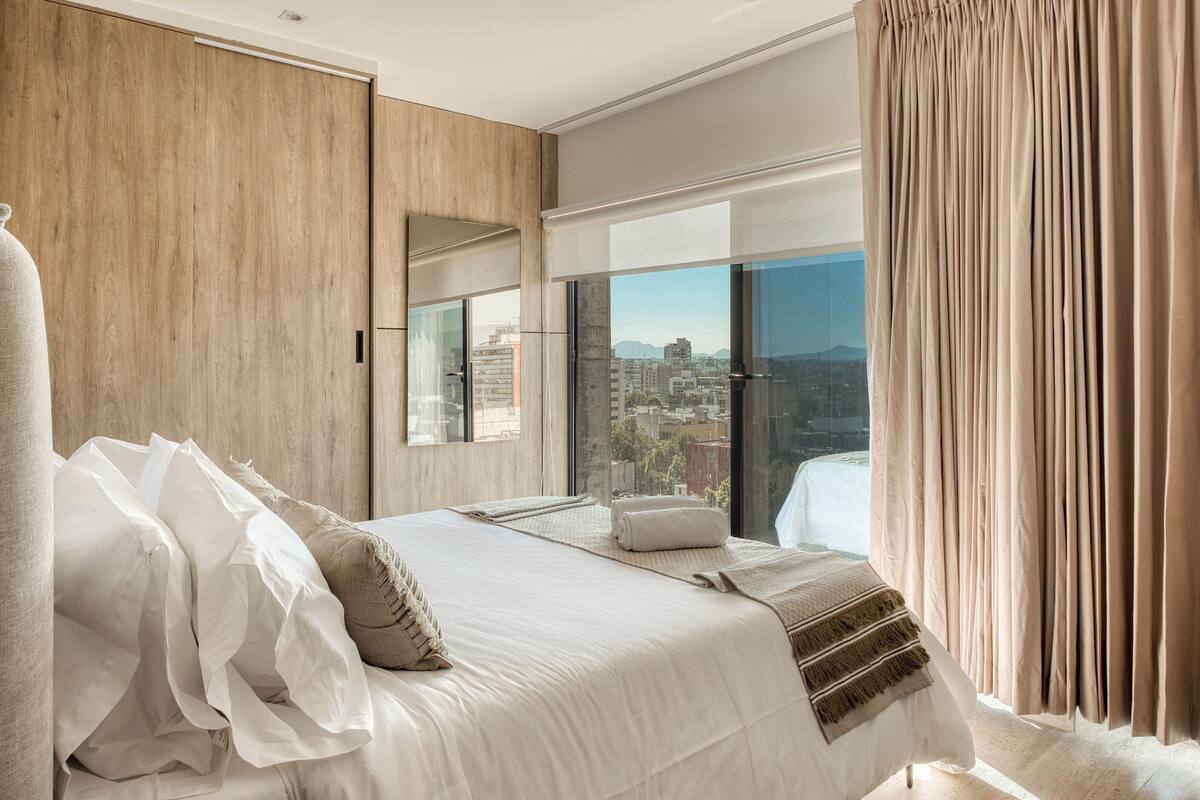 A modern hotel room with a large bed, a view of the city, and a wooden wardrobe.