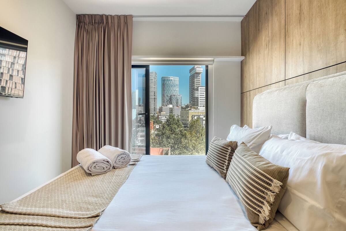 A modern hotel room with a large bed, a view of the cityscape, and a wooden accent wall.