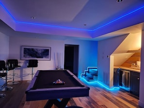 A modern living room with a pool table, bar stools, and a staircase leading to a kitchen area.