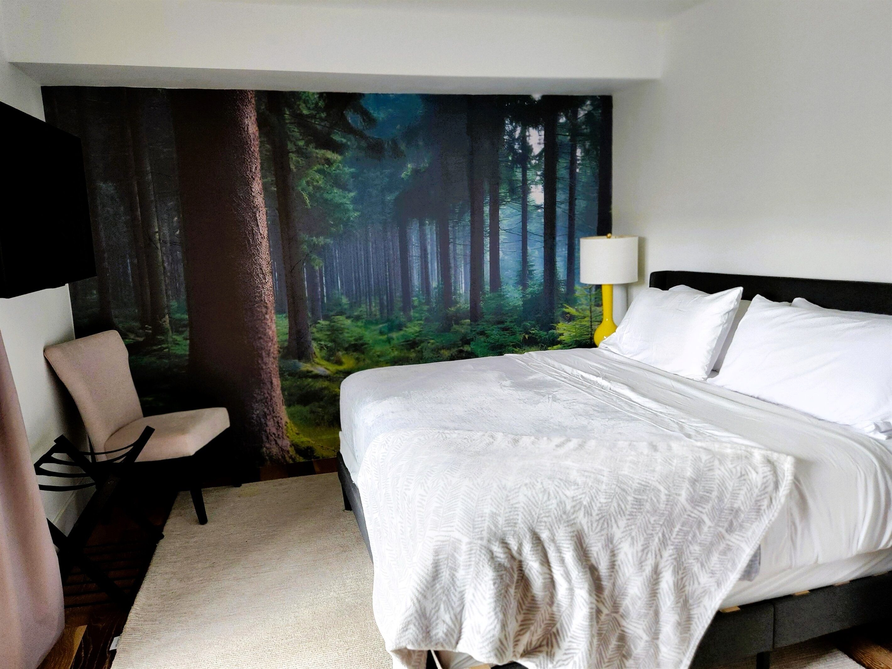 A bedroom with a bed, a chair, a wall mural of a forest, and a lamp.