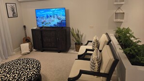 A living room with a flat-screen TV on a cabinet, a guitar, a patterned ottoman, and a plant.