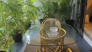 A glass-top table with a wicker chair in a covered outdoor area with potted plants.
