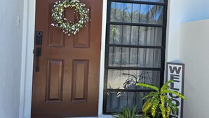 A brown door with a wreath, a welcome sign, a doormat, and two potted plants.