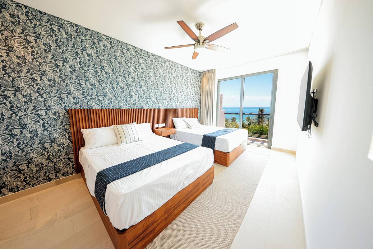 A hotel room with two beds, a ceiling fan, a large window with a view of the ocean, and a flat-screen TV mounted on the wall.