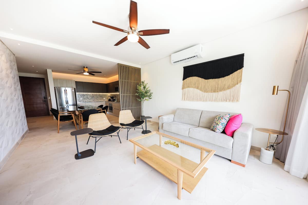 A modern living room with a sofa, coffee table, and chairs. There is a ceiling fan, an air conditioning unit, and a decorative wall hanging.
