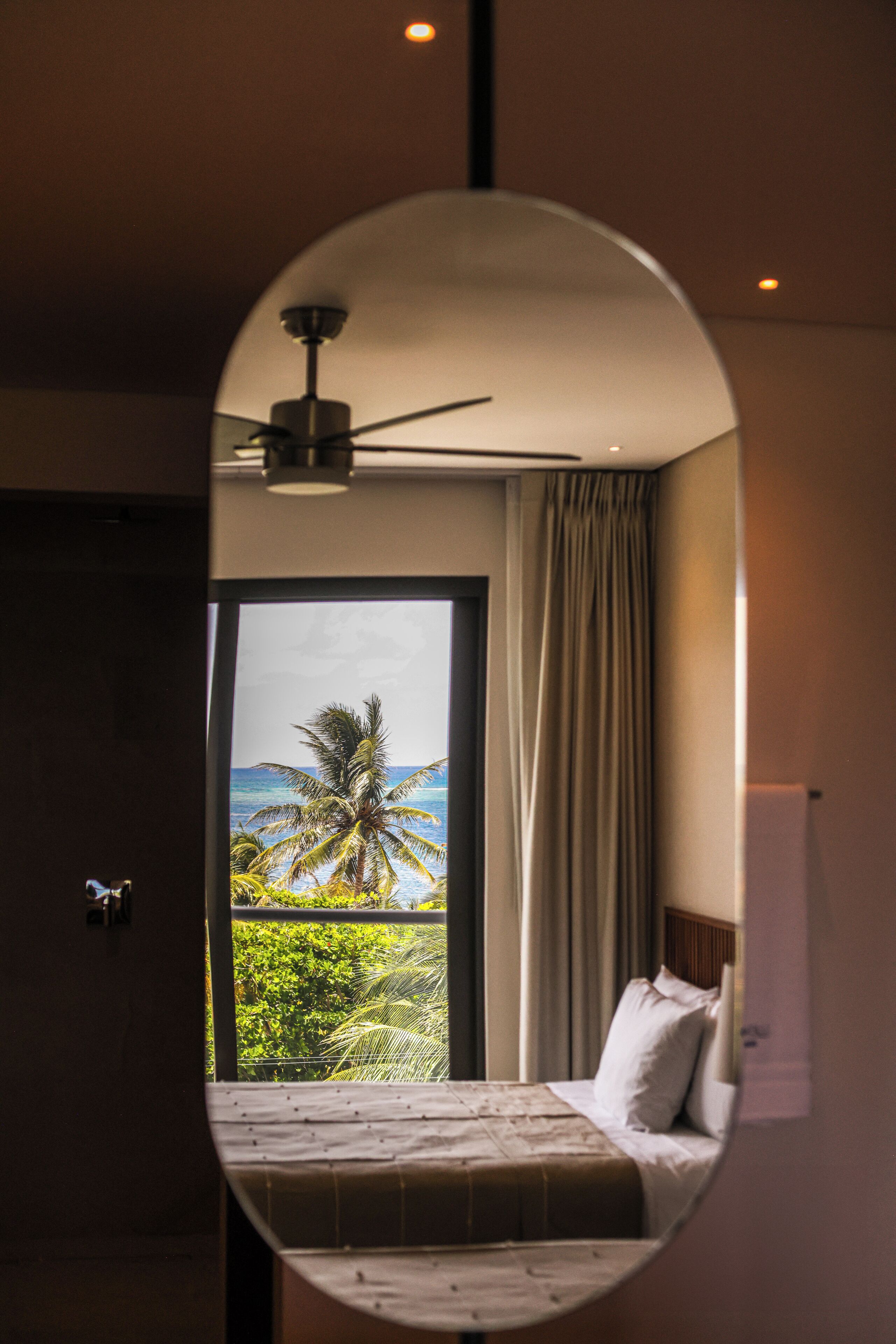 A bedroom with a bed, a ceiling fan, and a view of palm trees and the ocean through a large window.
