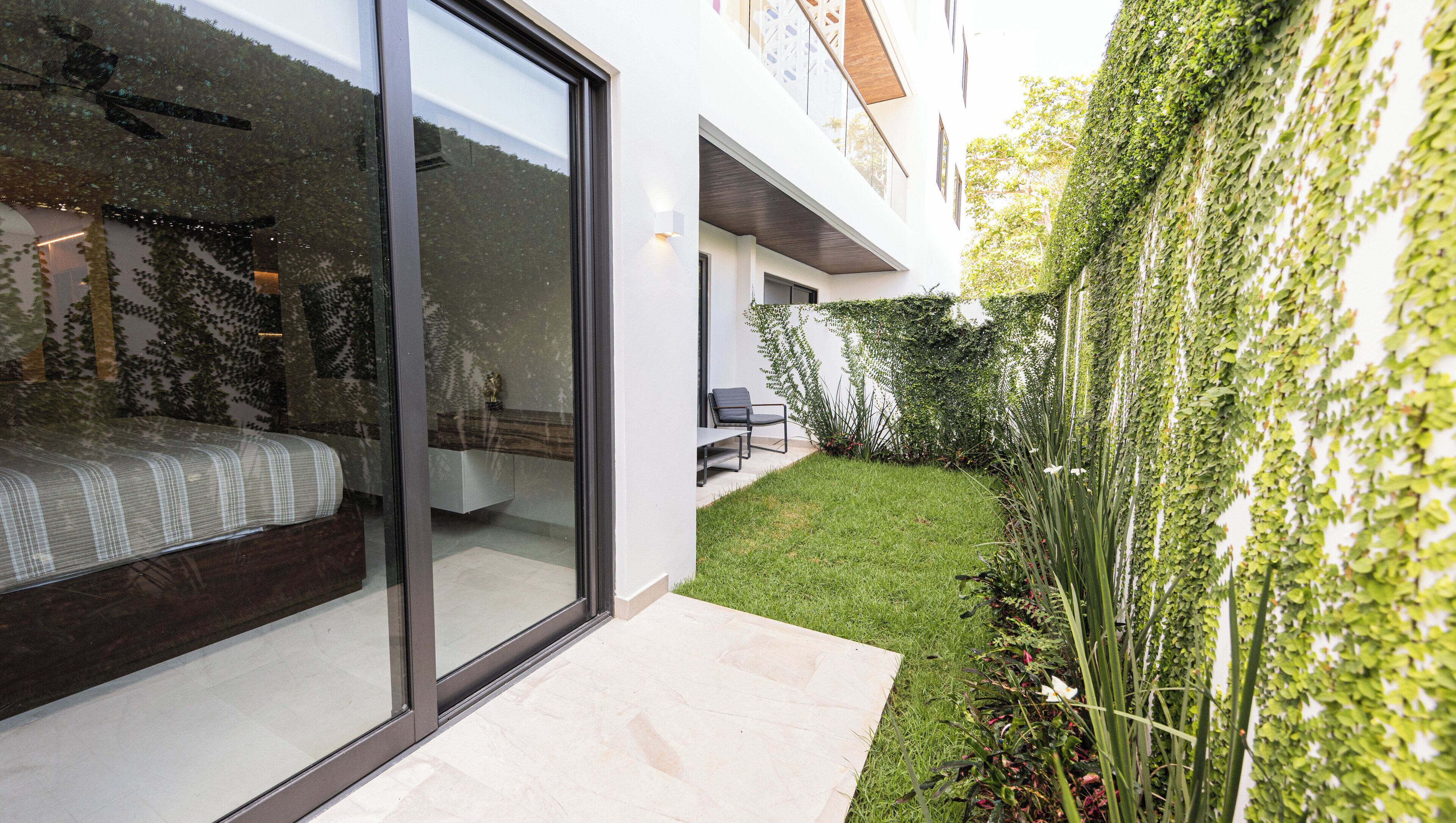 A modern house with a glass door leading to a bedroom, a small patio with a chair, and a lush green garden wall.