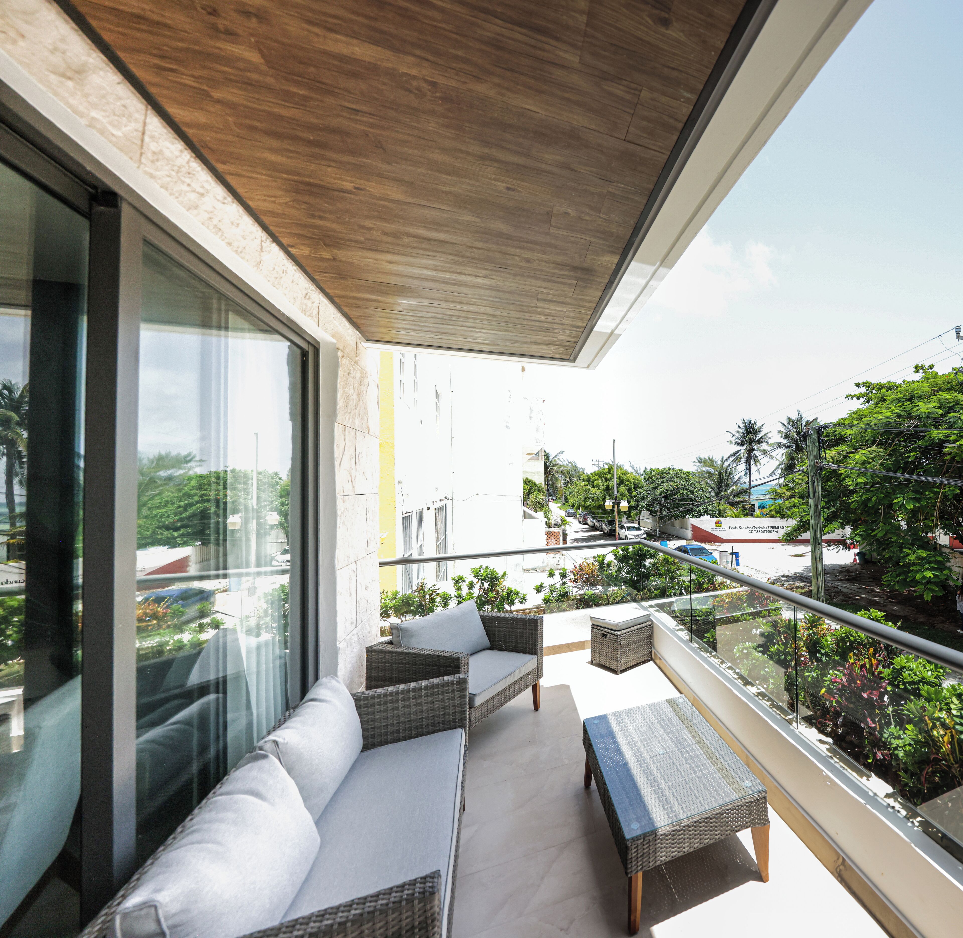 A balcony with wicker furniture, a glass table, and a view of the street and greenery.
