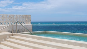A pool with steps leading to the water, a clear view of the ocean, and a white decorative wall.