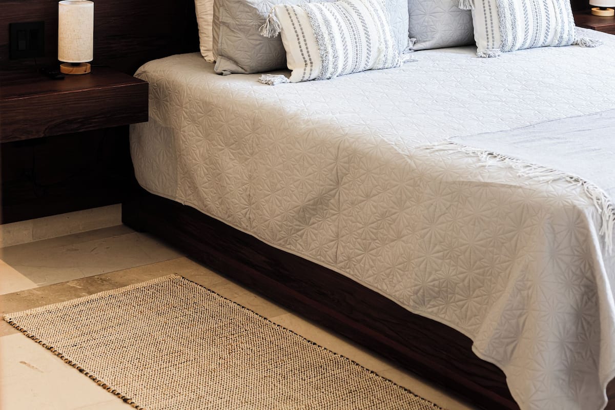 A neatly made bed with a light-colored quilt and multiple pillows, a wooden headboard, a bedside table with a lamp, and a woven rug on the floor.