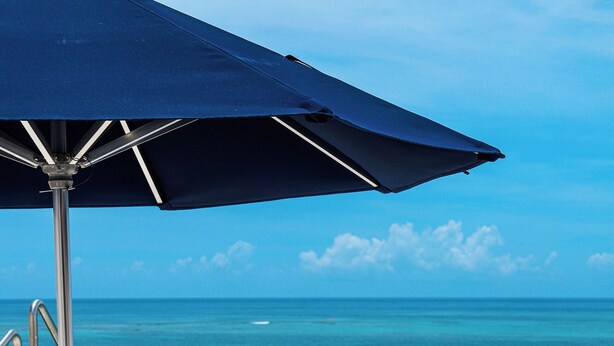 A lounge chair and small table under a blue umbrella, overlooking a pool and ocean.