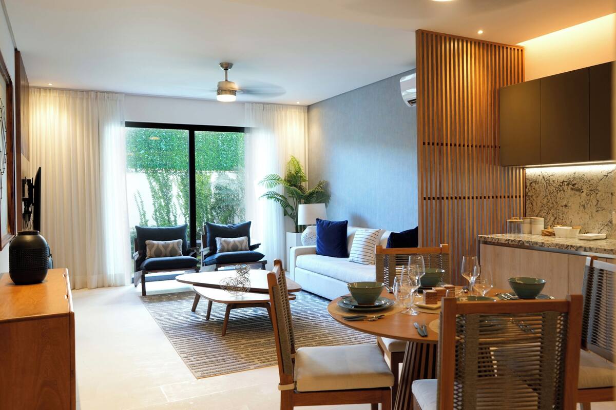A modern living room with a sofa, dining table, and chairs. There is a large window with curtains, a plant, and a TV mounted on the wall.