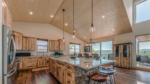 A spacious kitchen with wooden cabinets, a large island with stools, and a view of the outdoors. - Grindstone Cowboy | Luxury Lake Views, Hot Tub & Game Room Retreat (Ruidoso)