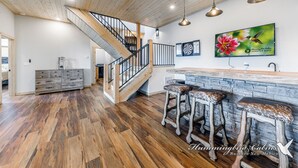A spacious living area with a wooden staircase, a stone bar counter, and a large TV mounted on the wall. - Grindstone Cowboy | Luxury Lake Views, Hot Tub & Game Room Retreat (Ruidoso)