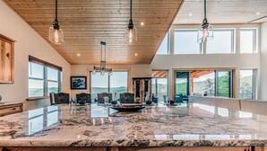 A spacious dining area with a large marble table, wooden ceiling, and large windows offering scenic views. - Grindstone Cowboy | Luxury Lake Views, Hot Tub & Game Room Retreat (Ruidoso)
