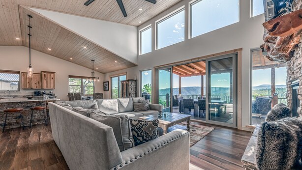 A spacious living area with a large gray sectional sofa, a stone fireplace, and a view of the outdoors through large sliding glass doors. - Grindstone Cowboy | Luxury Lake Views, Hot Tub & Game Room Retreat (Ruidoso)