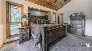 Une chambre avec un plafond en bois, un lit doté d’une tête de lit, une commode et une fenêtre donnant sur des arbres.
