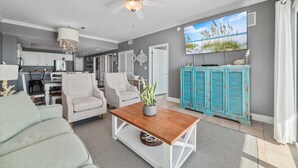 A spacious living room with a wooden coffee table, a ceiling fan, and a large window with a beach view. - Crystal Shores West 1003 | Indoor/Outdoor Pools, Fitness, Sauna & more | My Beach Getaways (Gulf Shores)