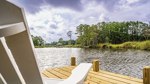 A wooden dock with a white chair facing a calm lake, surrounded by trees and a gazebo in the distance. - Charming 3-bedroom house on the water with a dock, close to Belhaven & Bath NC. (Belhaven)