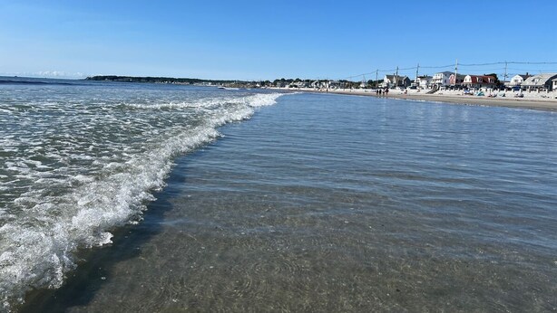 En strand med klart vand, sandet bredde og bygninger i baggrunden.