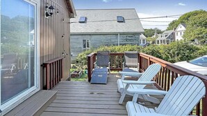 En træterrasse med hvide Adirondack-stole og et lille bord, med udsigt over et boligområde med huse og en klar himmel.