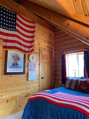 A wooden cabin room with a bed, an American flag, a framed picture, a clock, and a calendar.