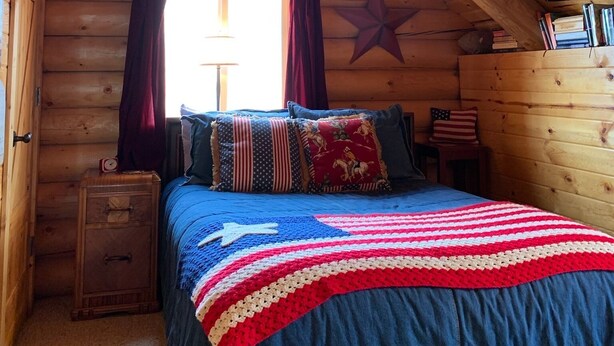 A cozy cabin room with a wooden interior, a bed with a red and white striped blanket, a star decoration on the wall, and an American flag. - Modern Log Cabin on a historic cattle ranch in Northern Nevada.  (Winnemucca)