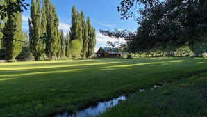 A large, open grassy field with a small house in the distance, surrounded by tall trees. - Modern Log Cabin on a historic cattle ranch in Northern Nevada.  (Winnemucca)