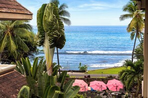 A view of the ocean from a balcony with large green plants and pink umbrellas. -  Ocean Views in Light & Bright 2 Bed condo across from Lyman's Cove- Kailua Kona (Kailua-Kona)