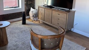A living room with a large flat-screen TV on a wooden cabinet, a wicker chair, a small round table, and a brick wall.