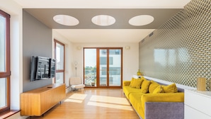 A modern living room with a wooden floor, a yellow sofa, a grey couch, a TV on a wooden cabinet, and a large window with a view of buildings.