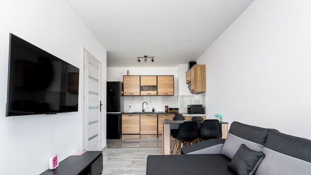 Ein modernes Wohnzimmer mit grauem Sofa, einem Flachbildfernseher und einer schlichten Küche im Hintergrund.
