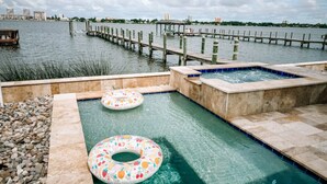 A swimming pool with two inflatable rings, a dock extending into a body of water, and a cityscape in the background.
